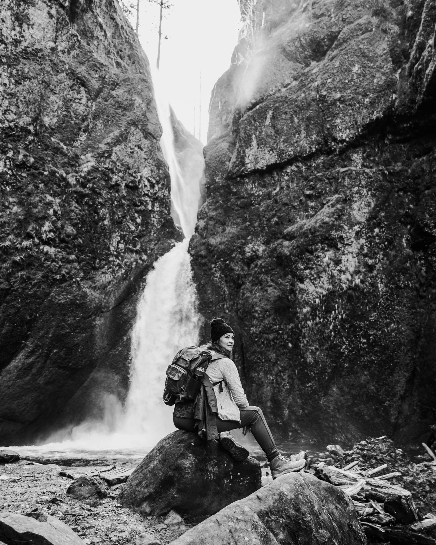 Black and white photo of a hiker with backpack sitting on rocks before a tall waterfall between rocky cliffs.