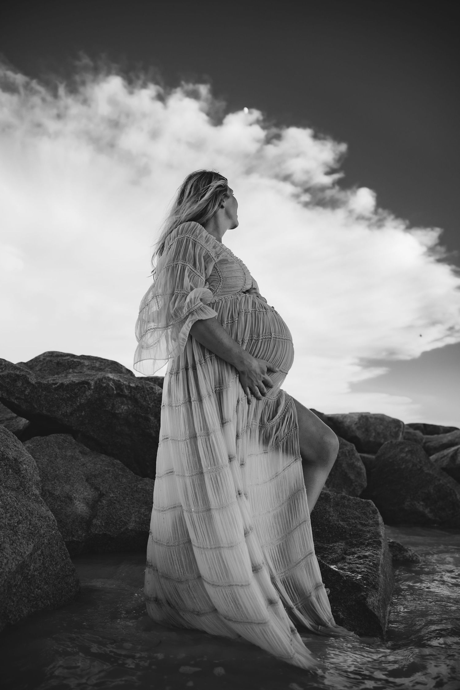 Black and white photo of pregnant woman in flowing dress standing on rocks by the ocean under a cloudy sky.