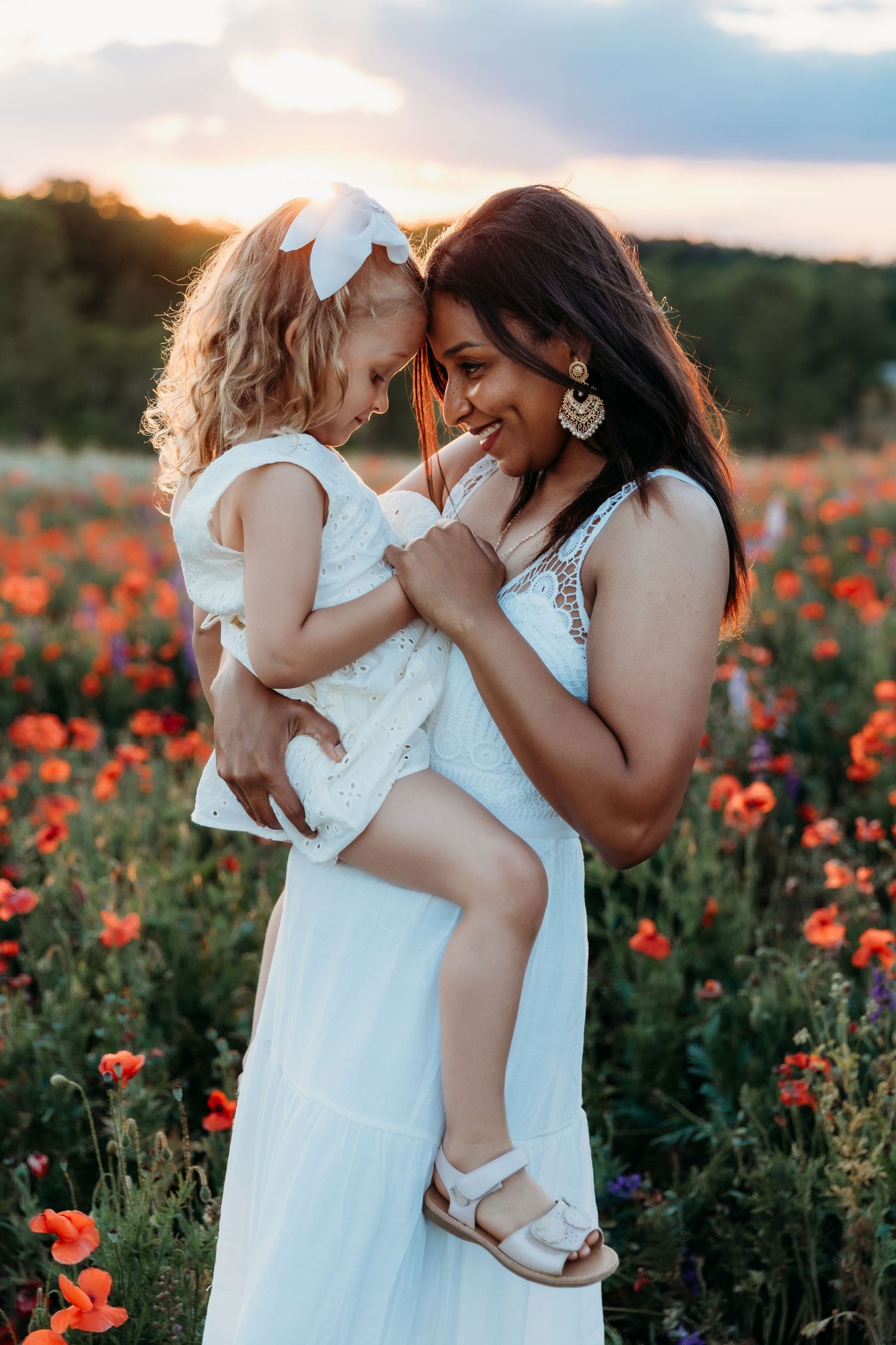 A mother in a white dress holds her toddler daughter in a blooming poppy field at sunset.