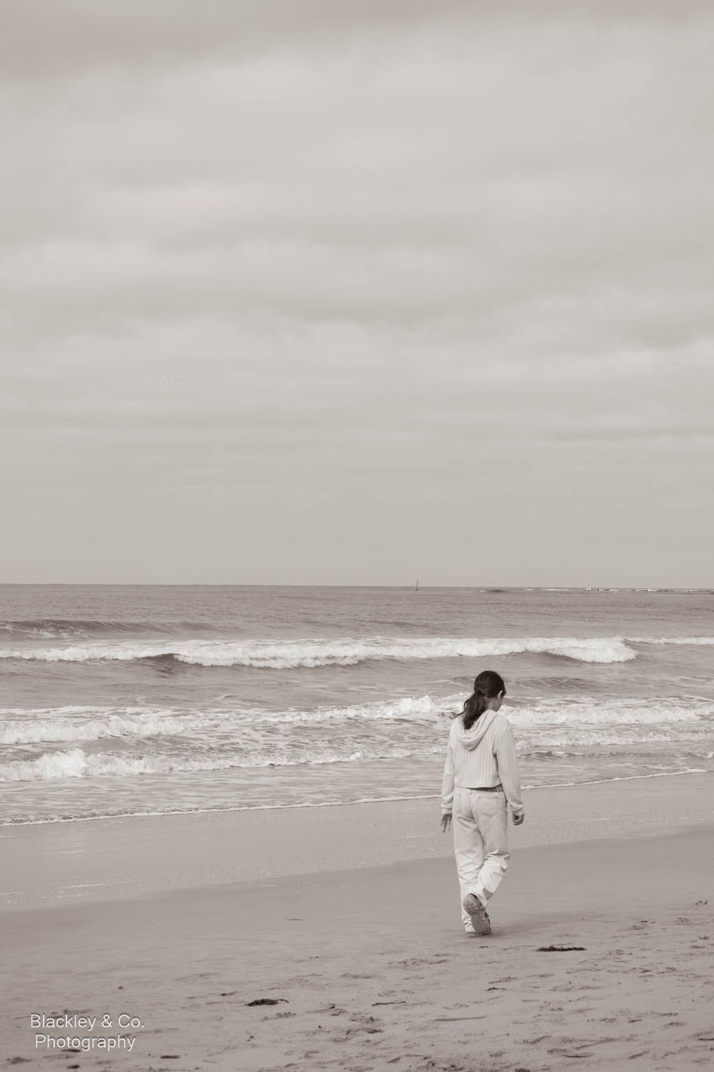 A lone woman in white walks along a serene beach shoreline in this moody black and white coastal photograph.