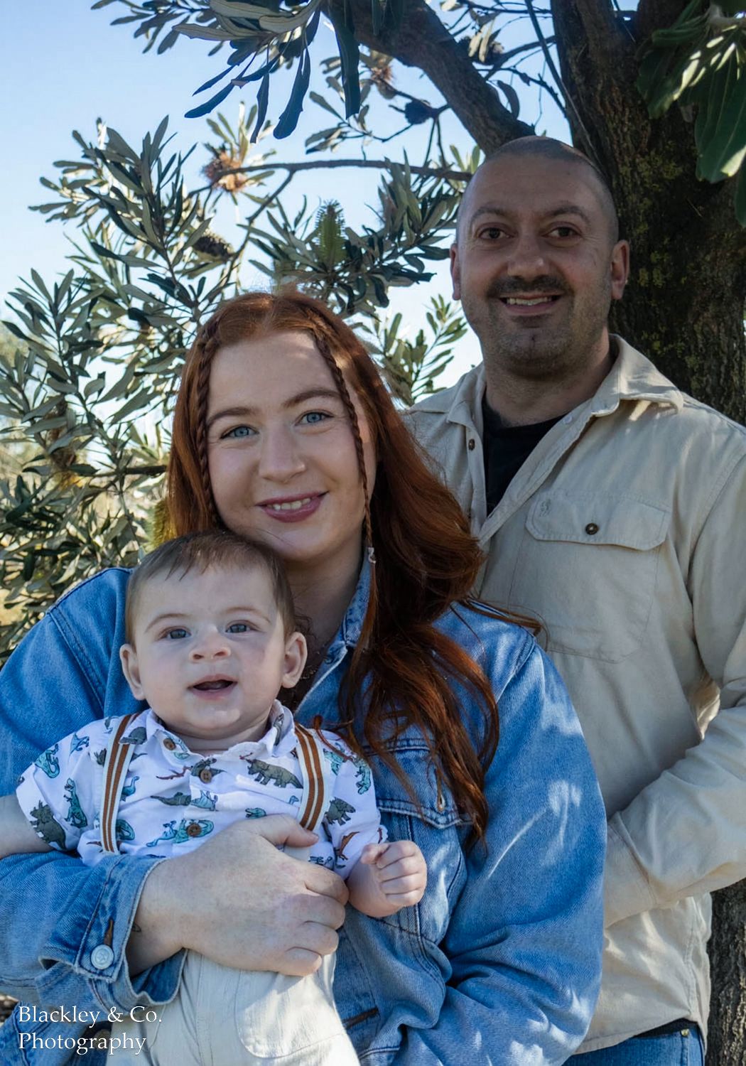 A young family with a redhead woman, bearded man, and smiling baby boy posing outdoors near a tree.