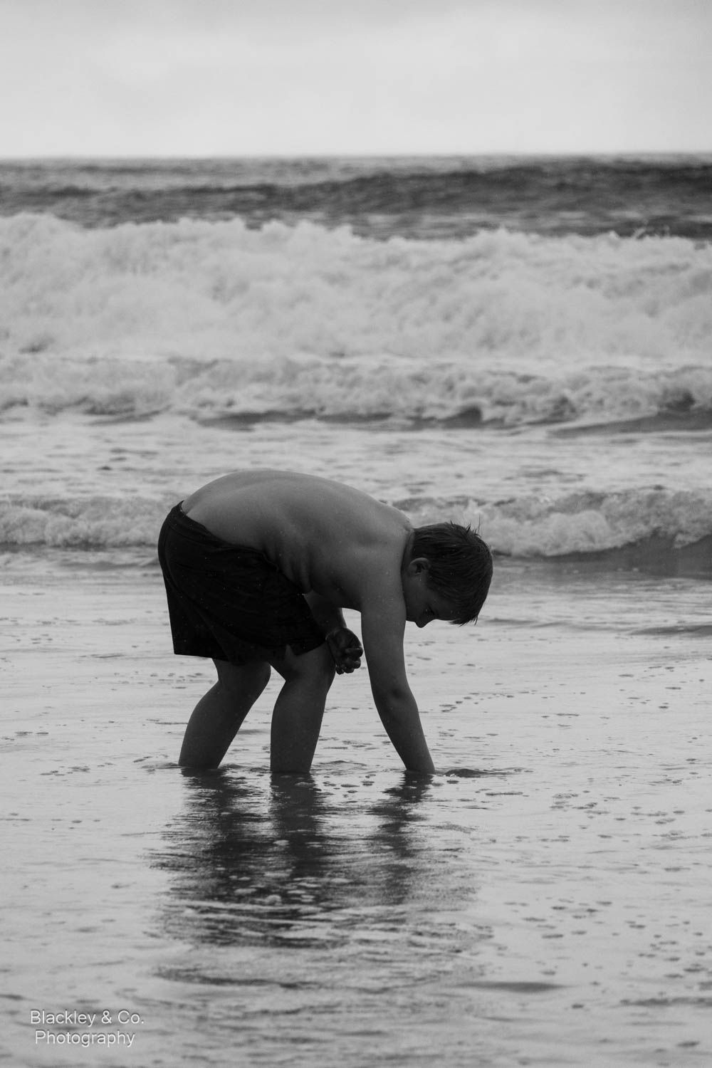Black and white photo of a shirtless boy bending down at the ocean shoreline with waves crashing behind him.