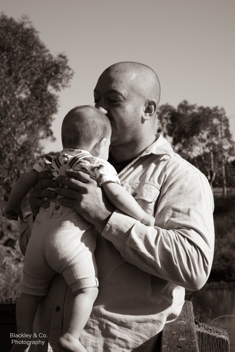Black and white photo of a bald man tenderly kissing his baby outdoors, captured by Blackley & Co. Photography.