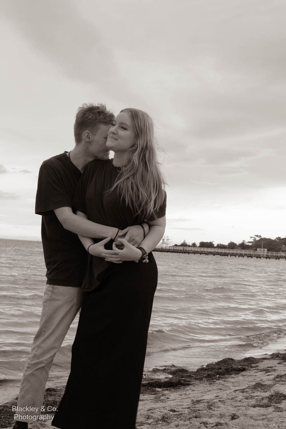 Black and white photo of a couple embracing and kissing on a beach with a pier in the background.
