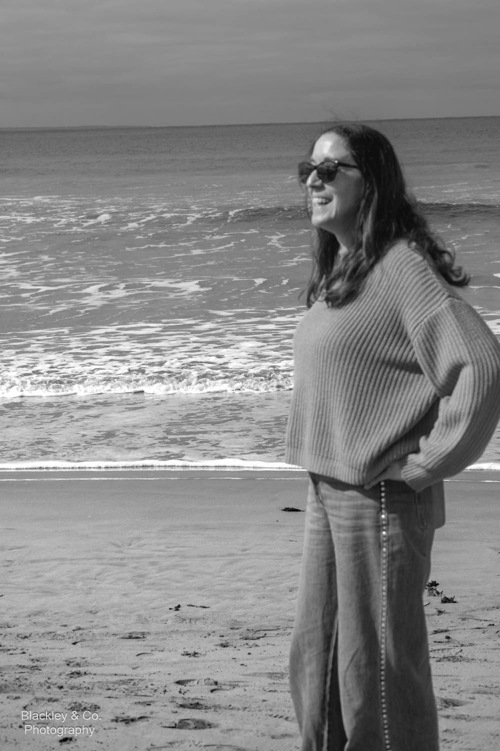 Black and white photo of a smiling woman in sunglasses and sweater standing on a beach with ocean waves behind her.