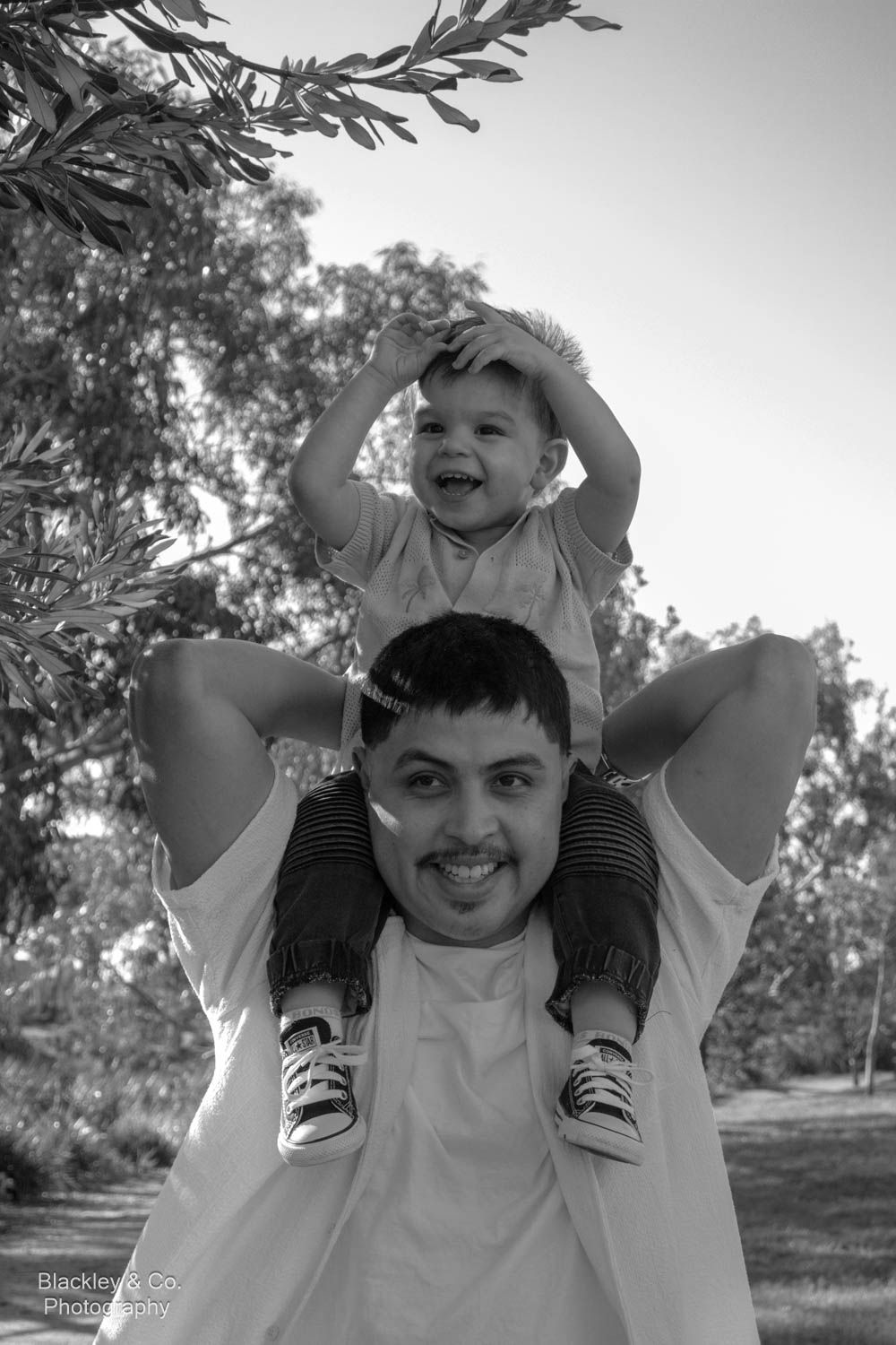 Black and white photo of a smiling man carrying a laughing toddler on his shoulders outdoors.