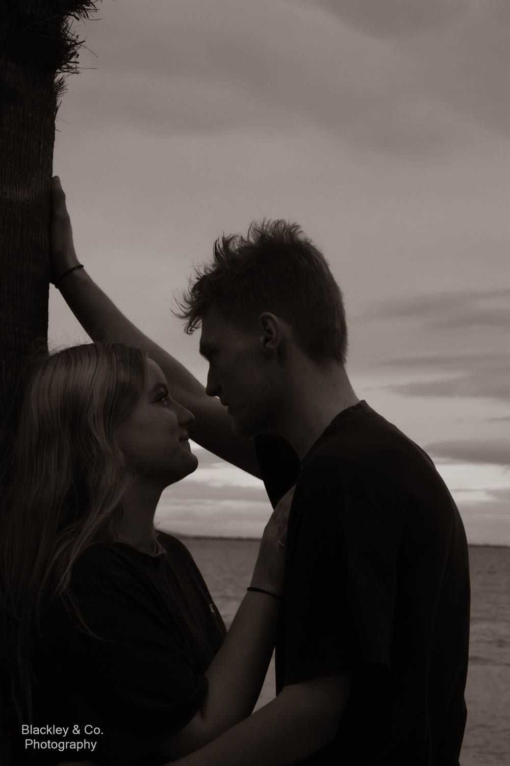 Black and white photo of a romantic couple leaning close together near a tree by the ocean at dusk.