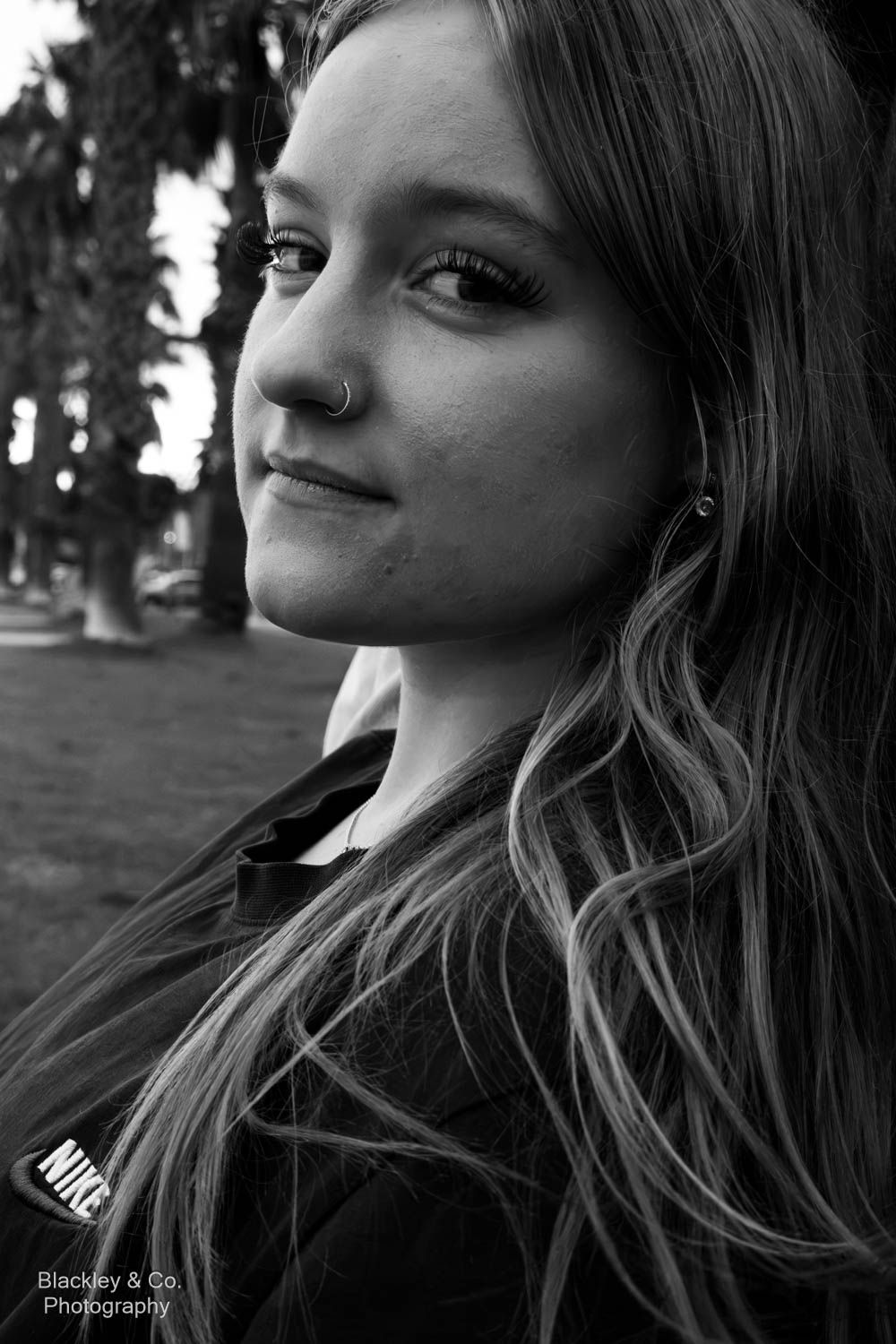 Black and white portrait of a young woman with long hair and nose ring, smiling softly outdoors near palm trees.