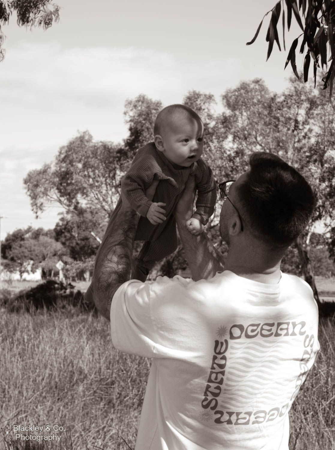Black and white photo of a man lifting a baby outdoors in a grassy field with trees in the background.