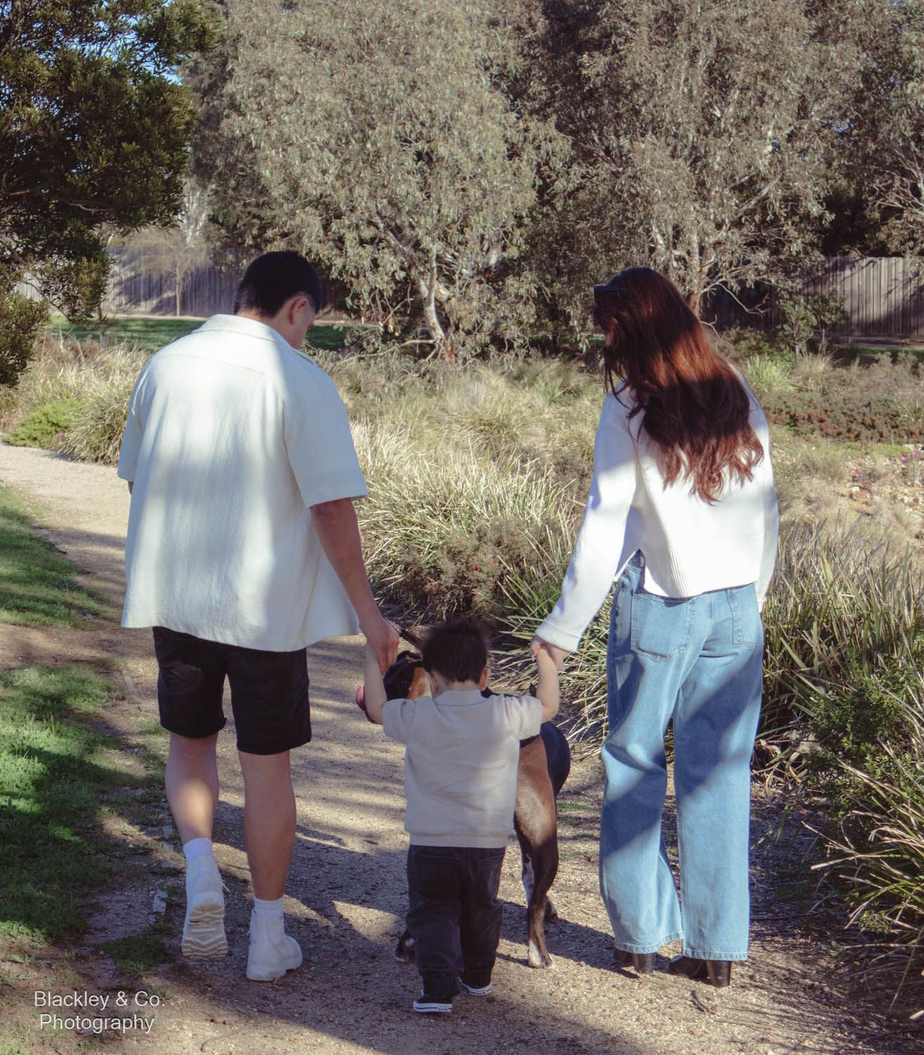 A family of three walks hand-in-hand down a sunny park path, parents guiding their toddler through lush greenery.