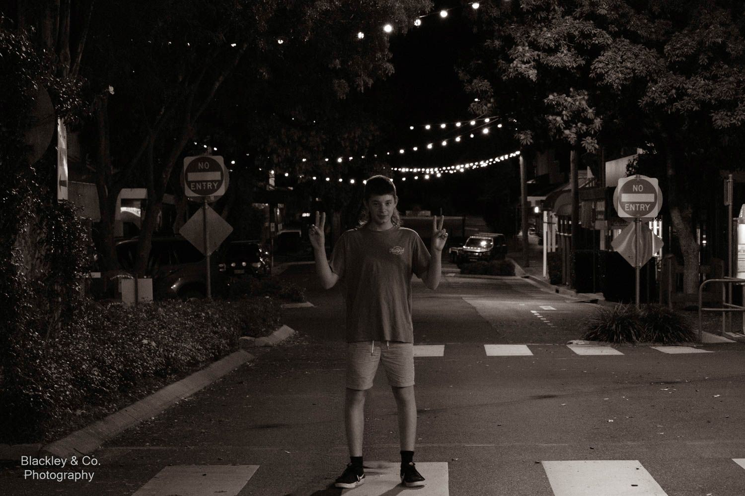 Man standing on crosswalk at night with string lights on tree-lined street, black and white photography.