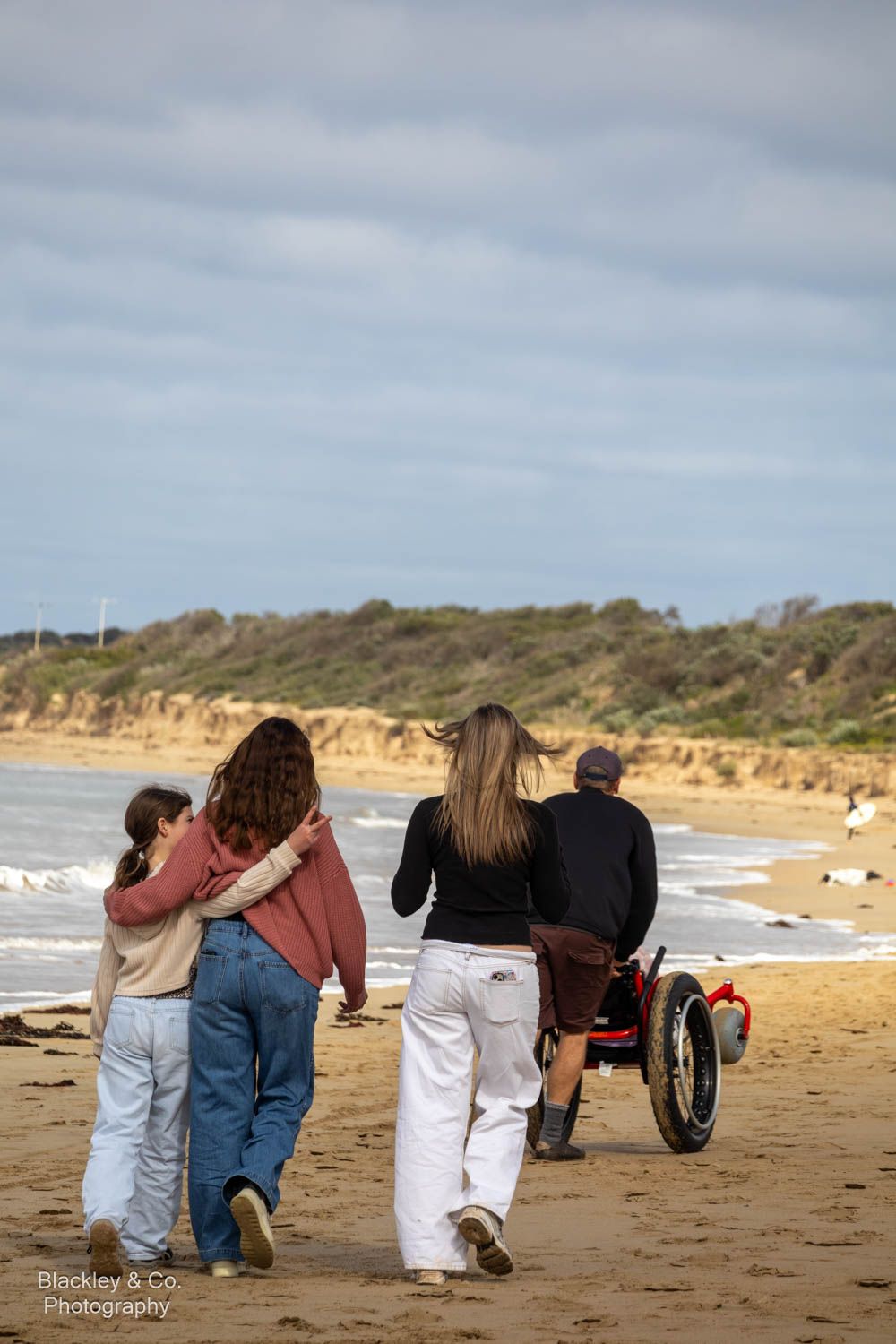 Four people walking on a sandy beach together, one using a wheelchair, with ocean waves and green dunes in the background.