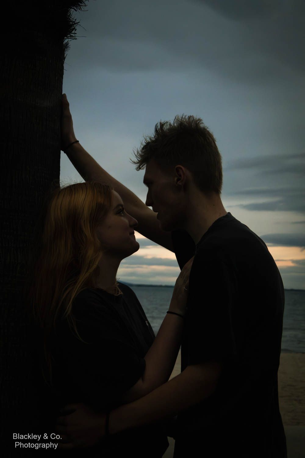 Couple sharing a romantic moment at sunset on a beach, silhouetted against a dramatic cloudy sky.