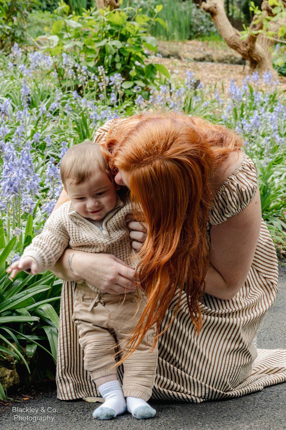Red-haired mother kissing smiling baby in beige knit outfit among purple bluebells in garden.