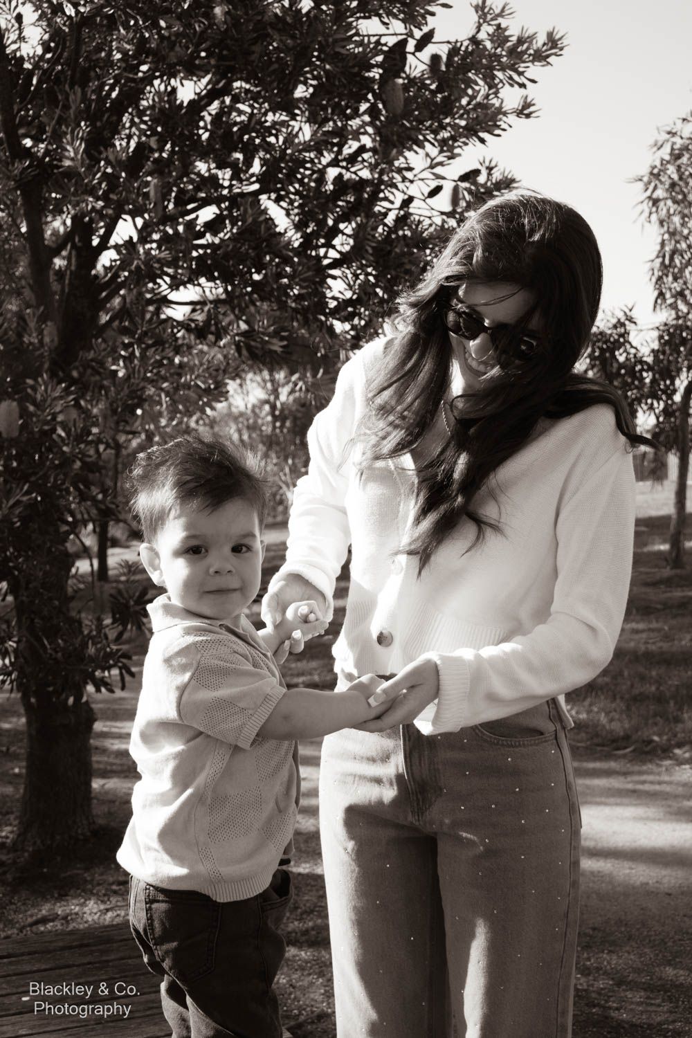 Black and white photo of a young boy and woman in sunglasses sharing a moment outdoors near trees.