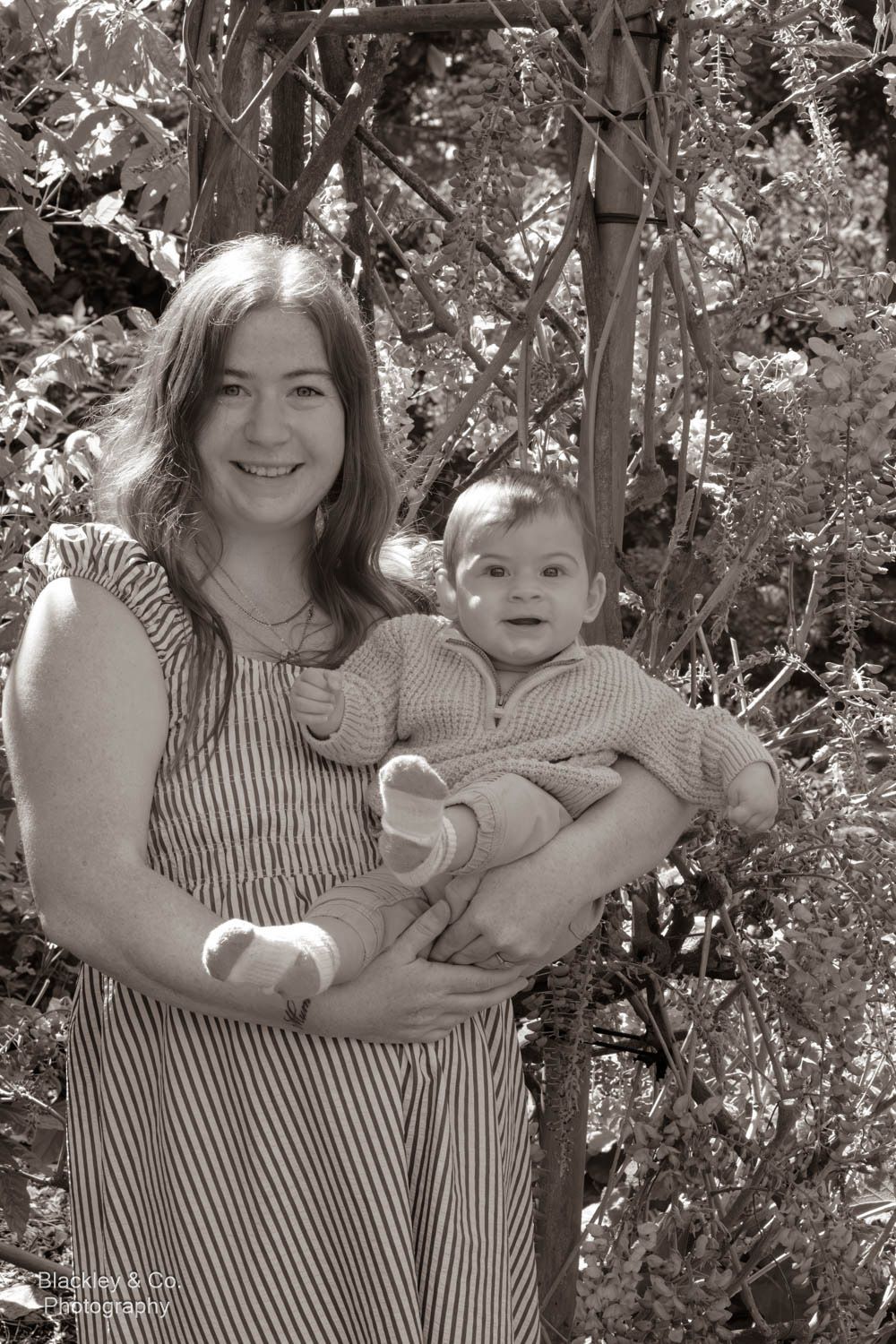 Black and white photo of a smiling young woman holding a happy baby outdoors in a garden setting.