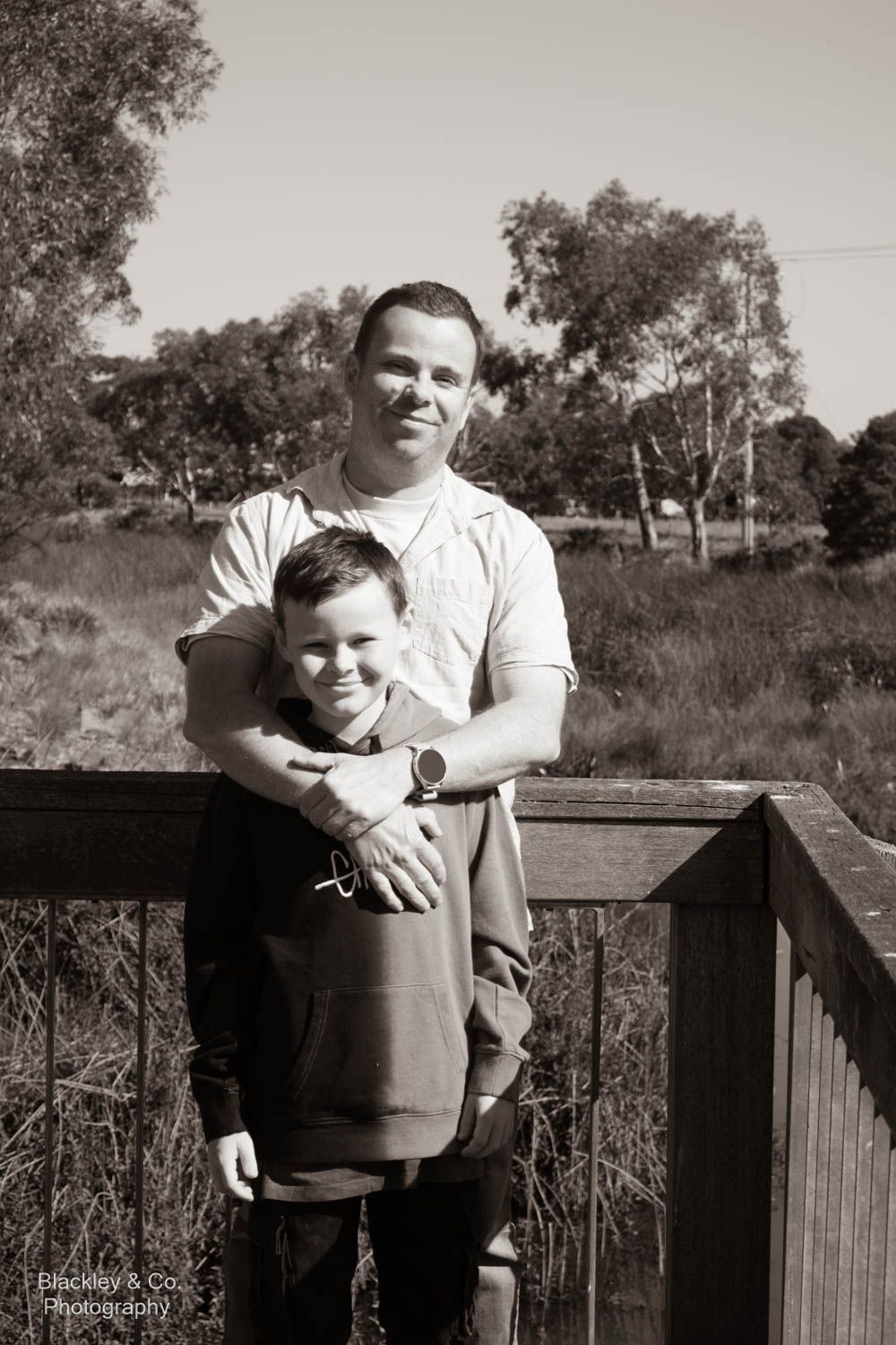 Black and white photo of a man and young boy posing together on a wooden deck surrounded by Australian bushland.