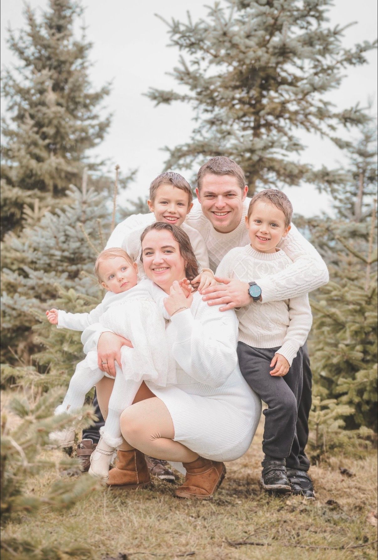 Happy family of five posing together among pine trees in matching white sweaters.