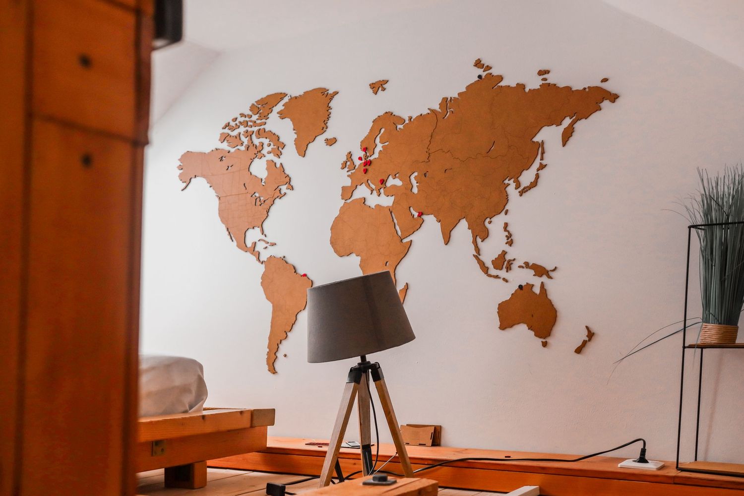 Decorative wooden world map wall art above a lamp on a wooden tripod base in a bedroom.