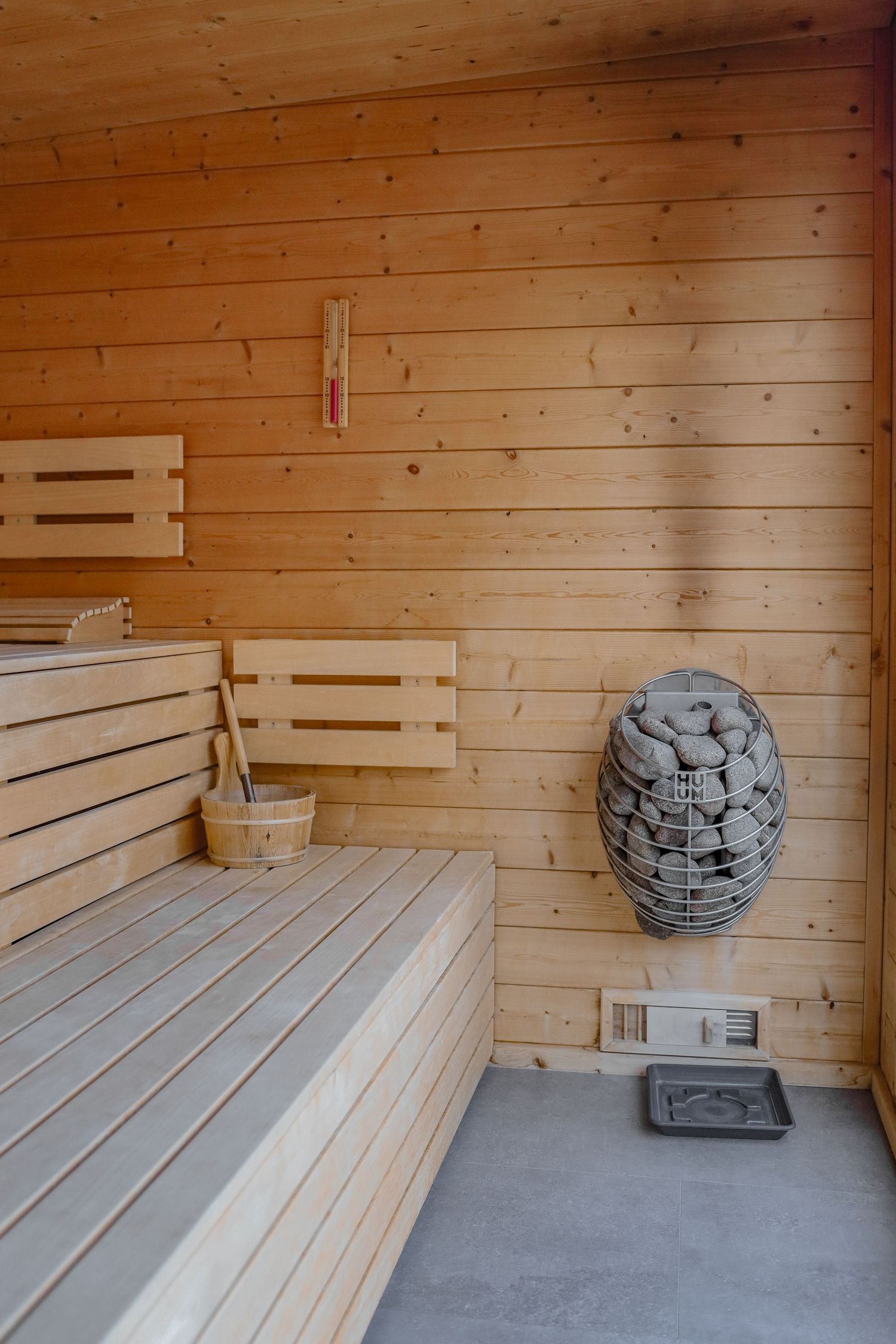 Interior of a modern wooden sauna with benches, bucket, and HUUM stone heater.
