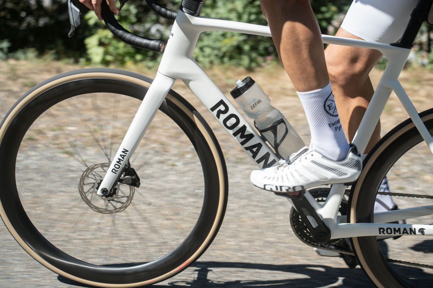 Cyclist riding a white Roman road bike on cobblestones, wearing white cycling shoes and socks.