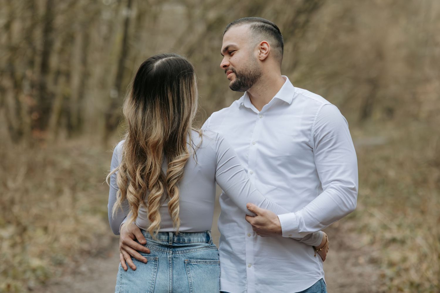 Romantic couple in white shirts embracing on a forest path surrounded by bare trees.