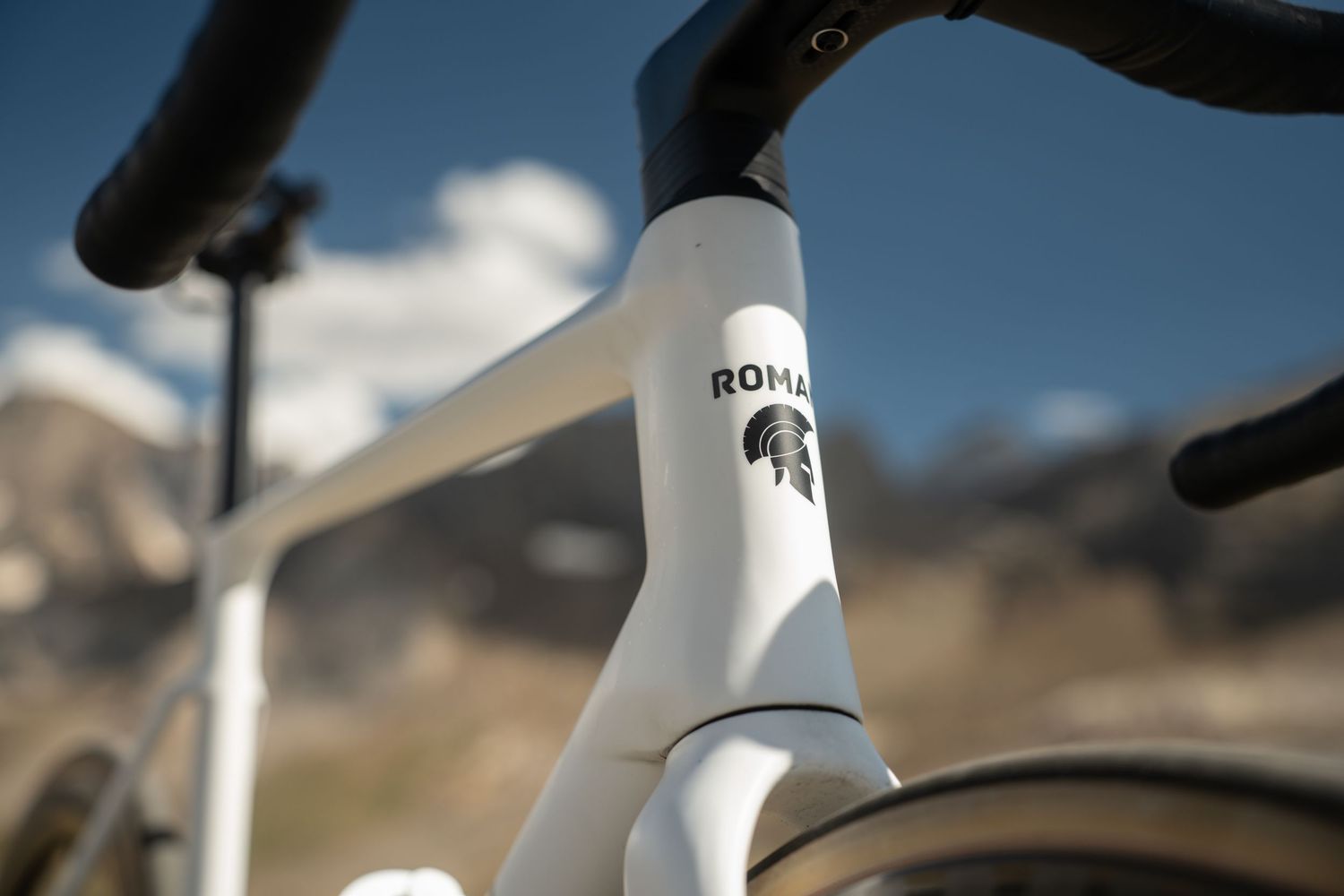 Close-up of a white Roman branded road bike frame with a Spartan helmet logo against a mountain backdrop.