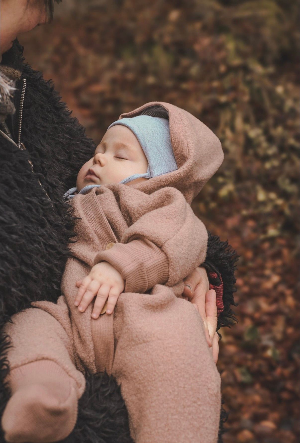 Sleeping newborn baby in a pink fleece suit with blue bonnet held by a parent outdoors.