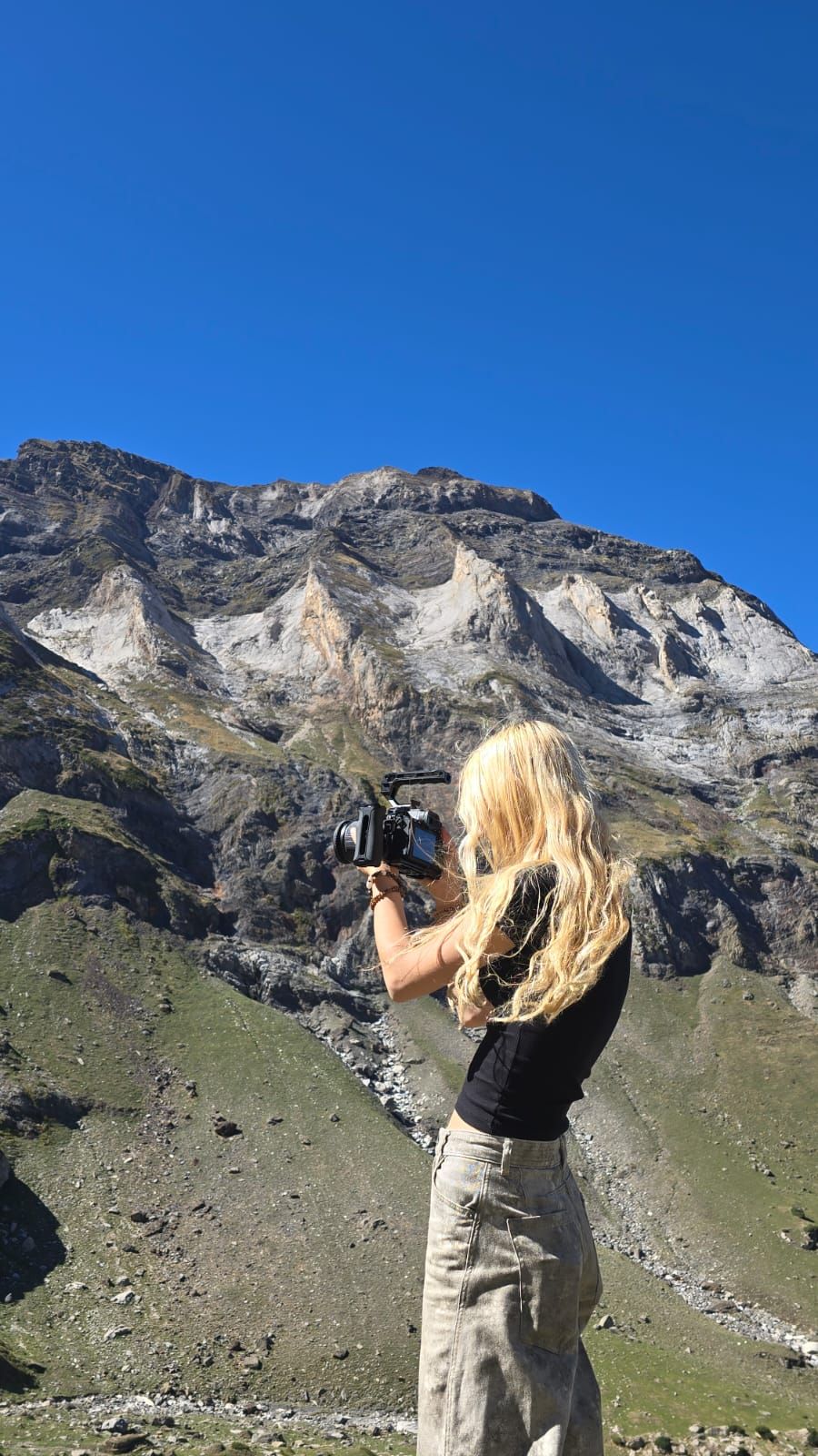 Blonde woman photographing a rocky mountain landscape under a clear blue sky.