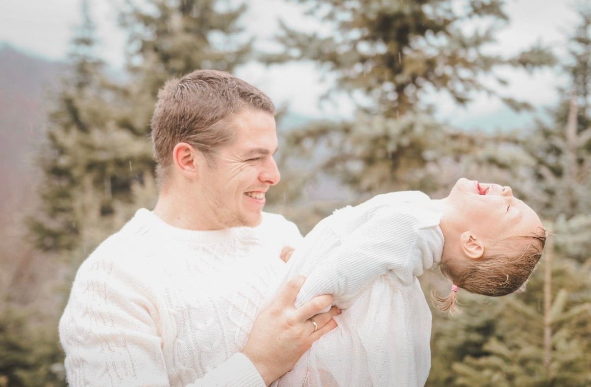Smiling father holding a laughing baby outdoors with evergreen trees in the background.