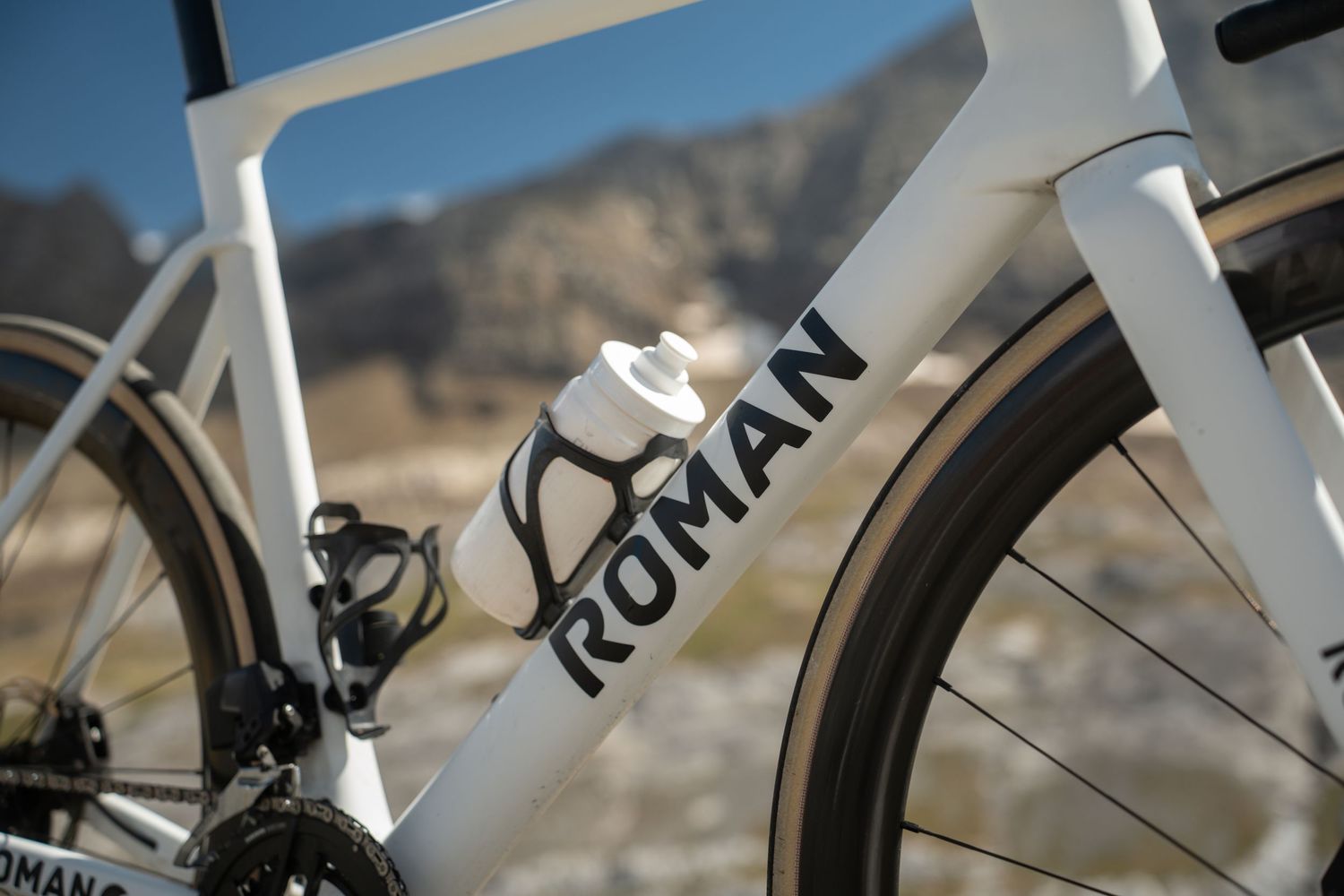 Close-up of a white Roman road bike frame with water bottle and mountain backdrop.