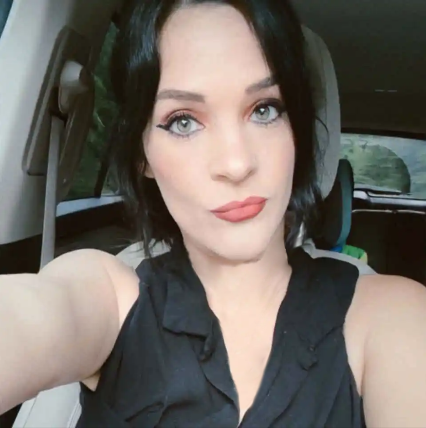 Young woman with dark hair and blue eyes taking a selfie in a car, wearing a black top with coral lipstick.
