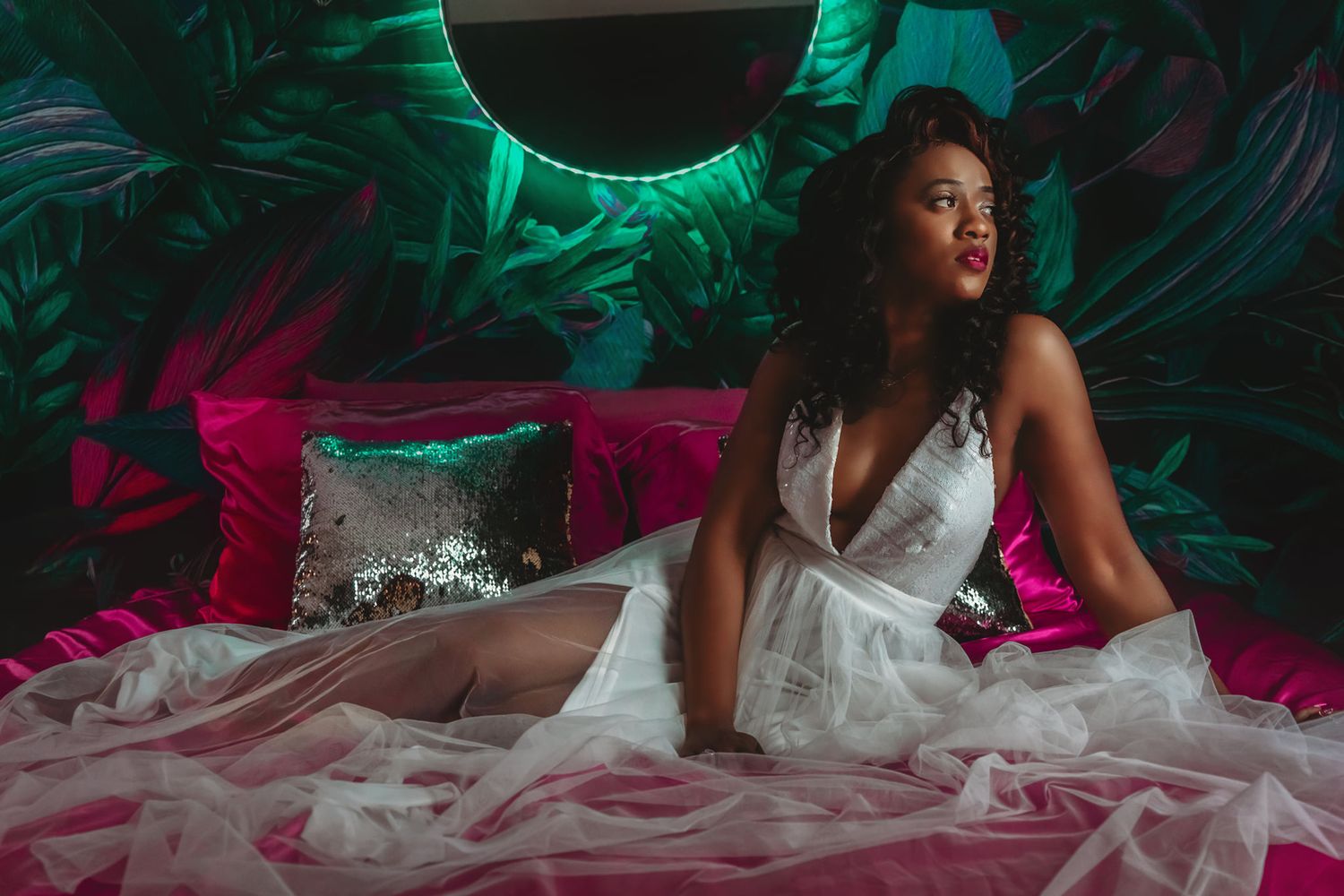 Woman in white lingerie sitting on pink bed in tropical neon-lit boudoir setting.