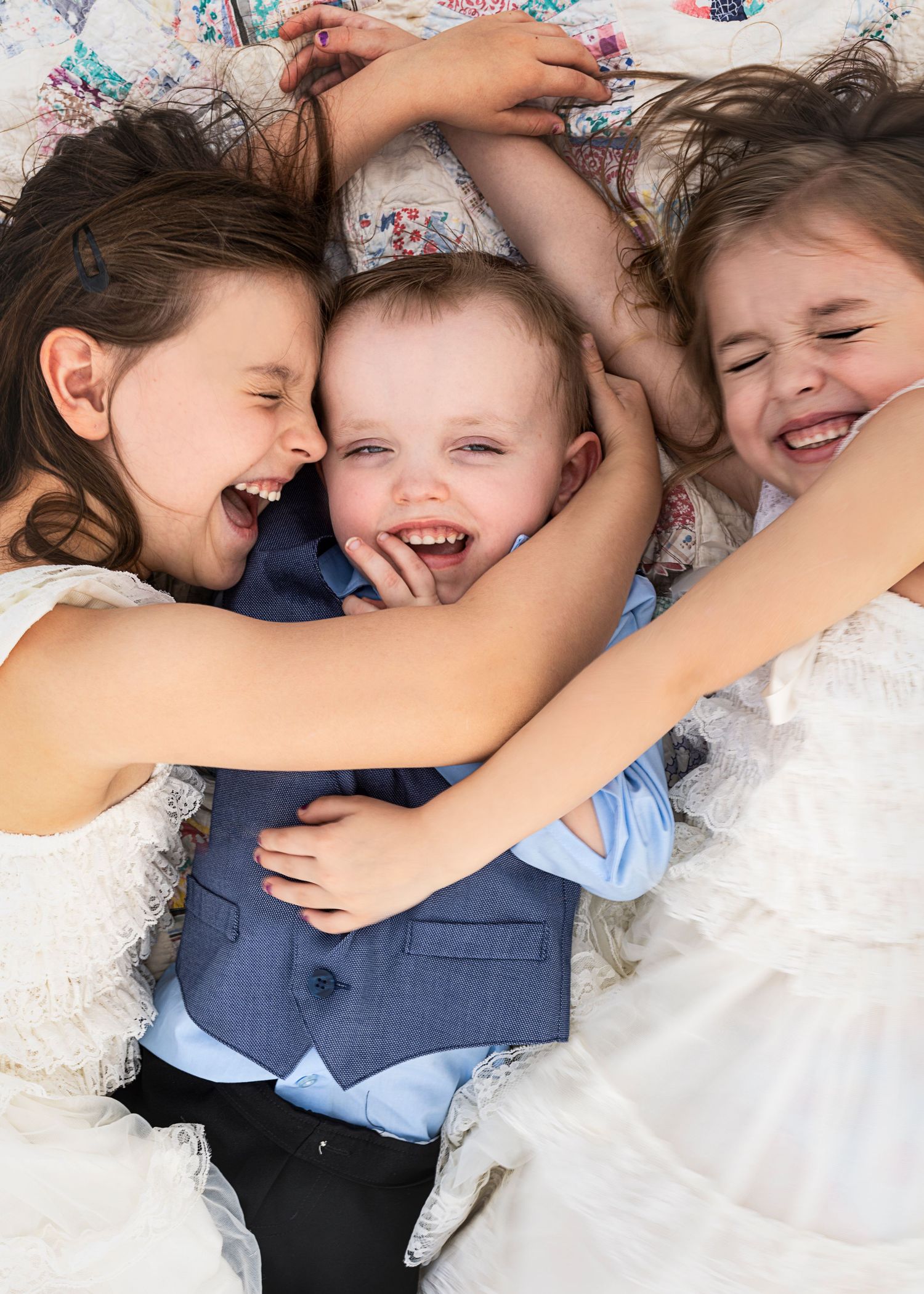 Three joyful children in formal wear laugh and hug while lying on a floral quilt.