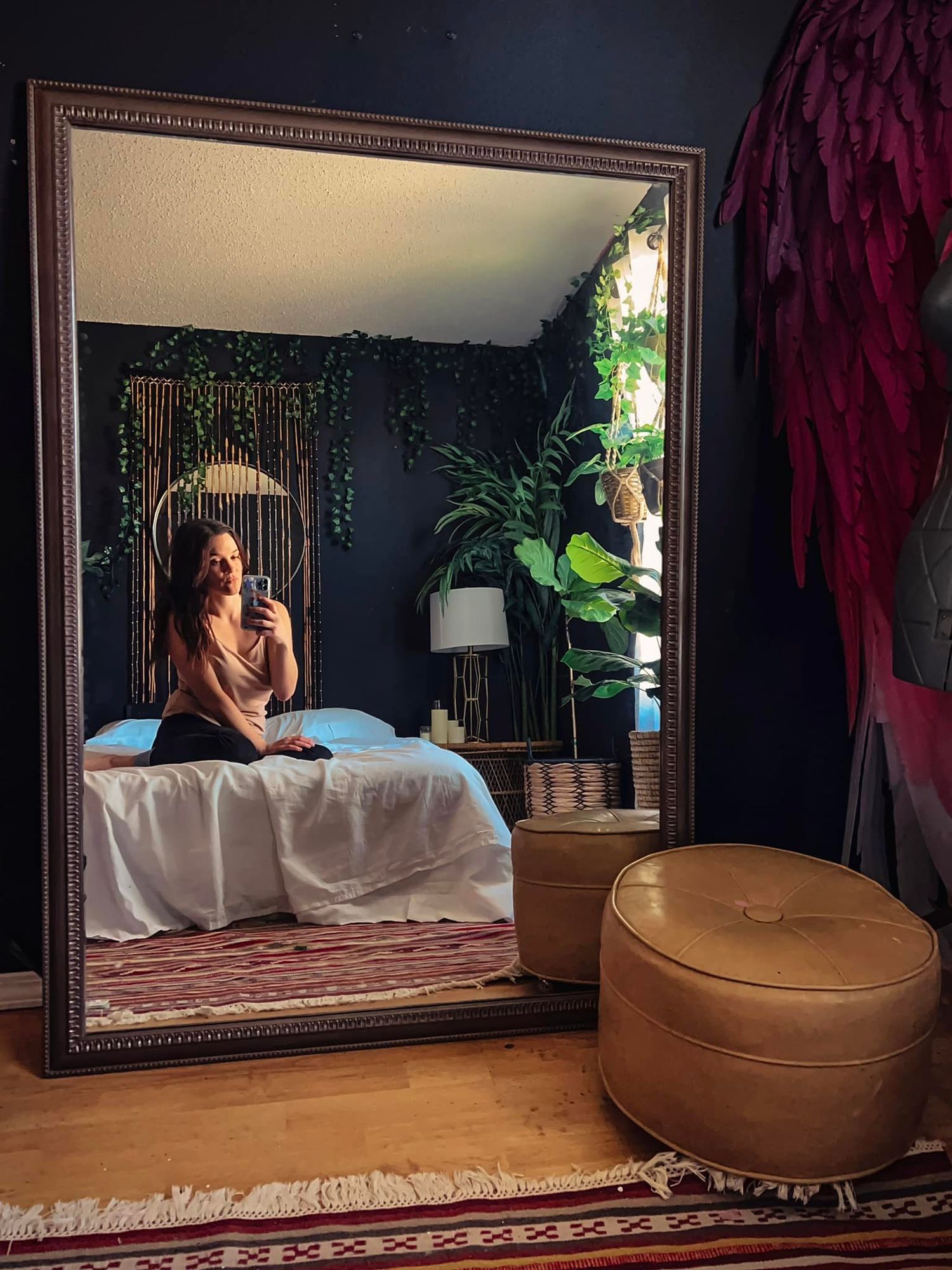 Woman takes mirror selfie in bohemian bedroom decorated with plants and large ornate mirror.