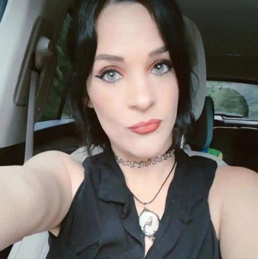 Woman with black hair and blue eyes taking a selfie in a car, wearing a black top and choker necklace.