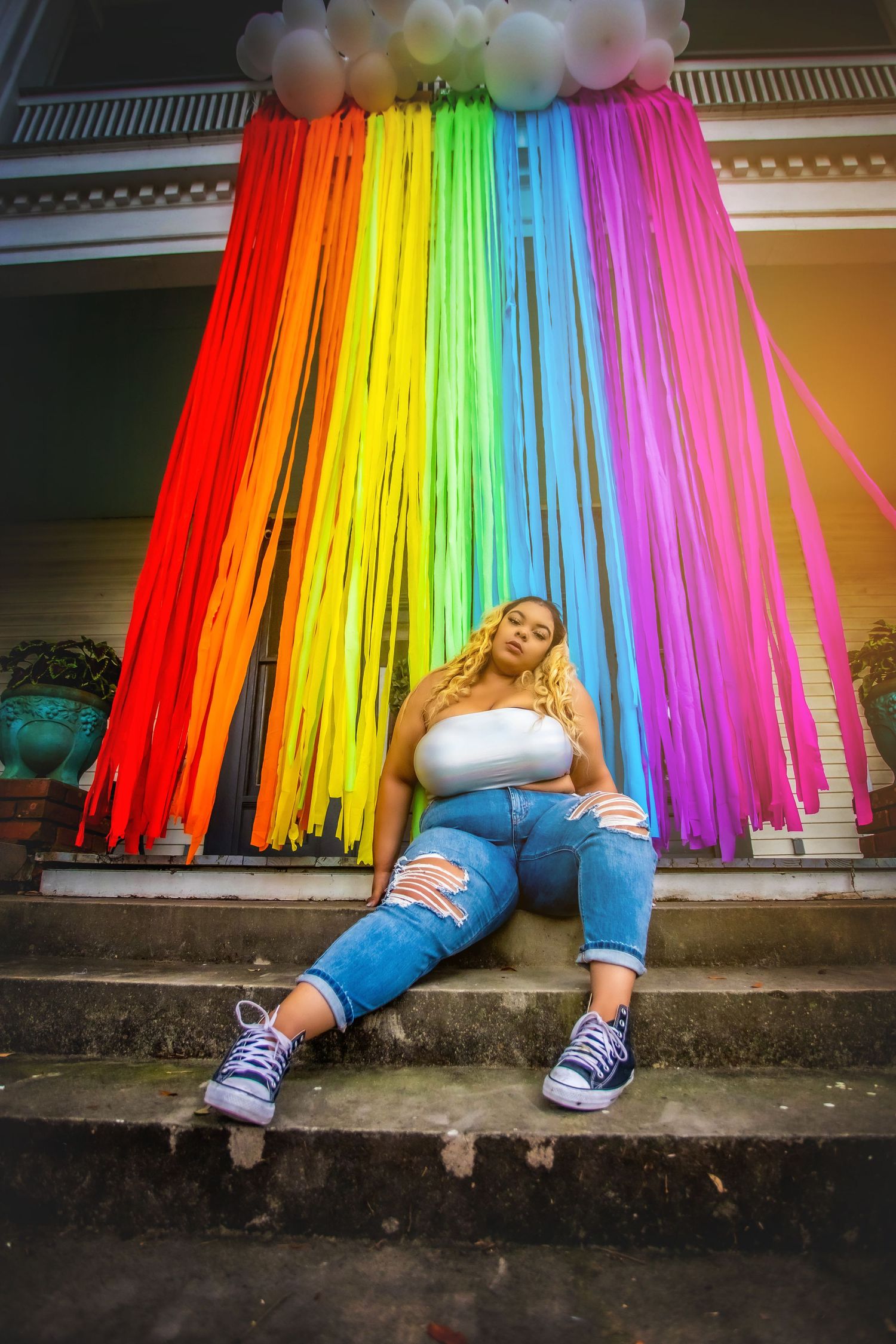 Plus-size woman in white top and ripped jeans relaxes on steps before vibrant rainbow streamer backdrop.