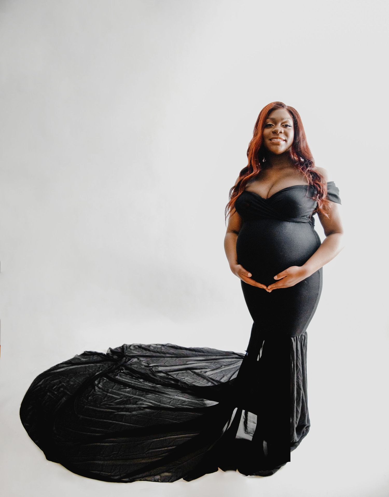 Pregnant woman in elegant black off-shoulder mermaid gown with flowing train on white background.
