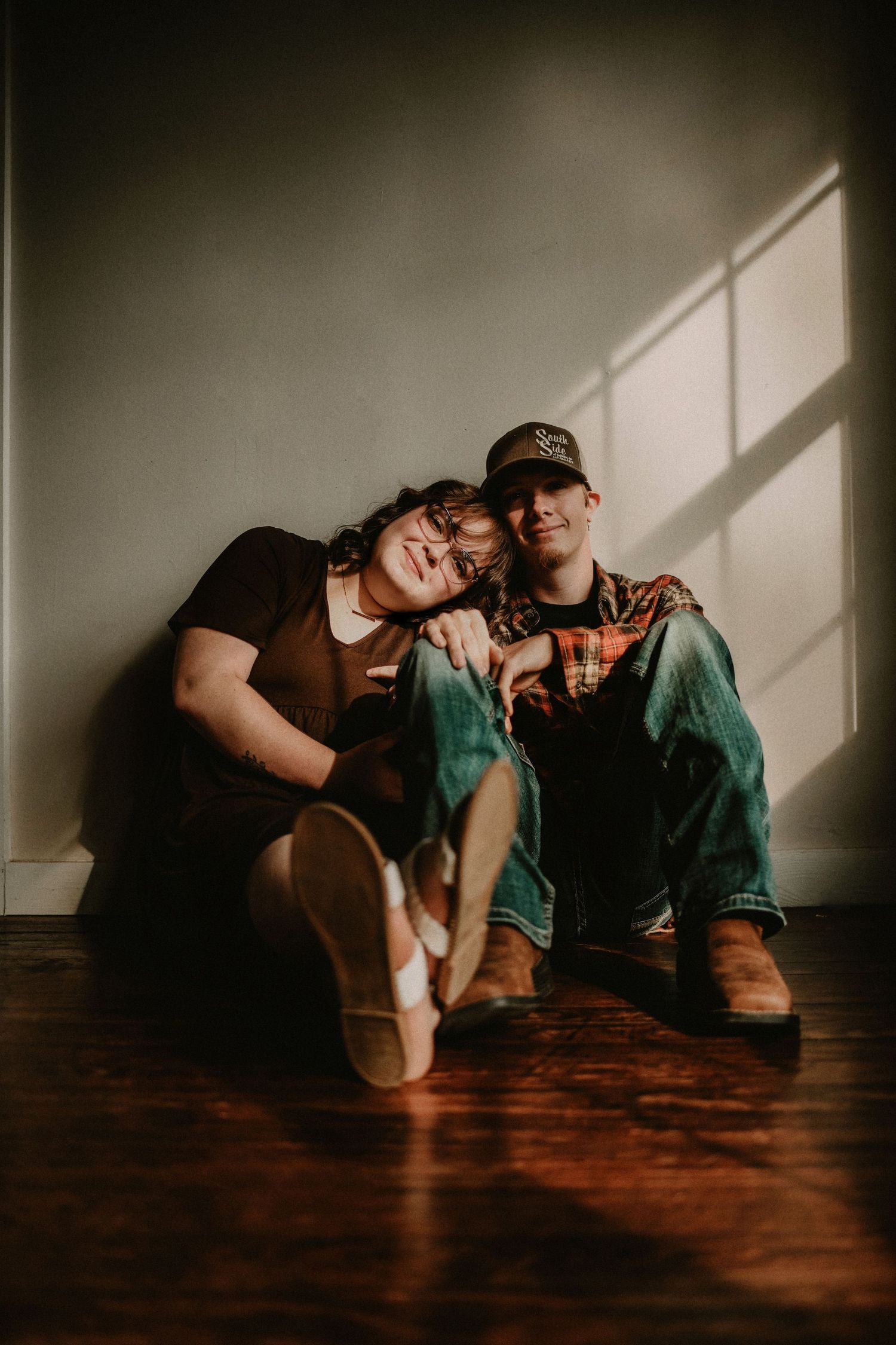 Young couple sitting on hardwood floor leaning together in warm sunlight against gray wall.