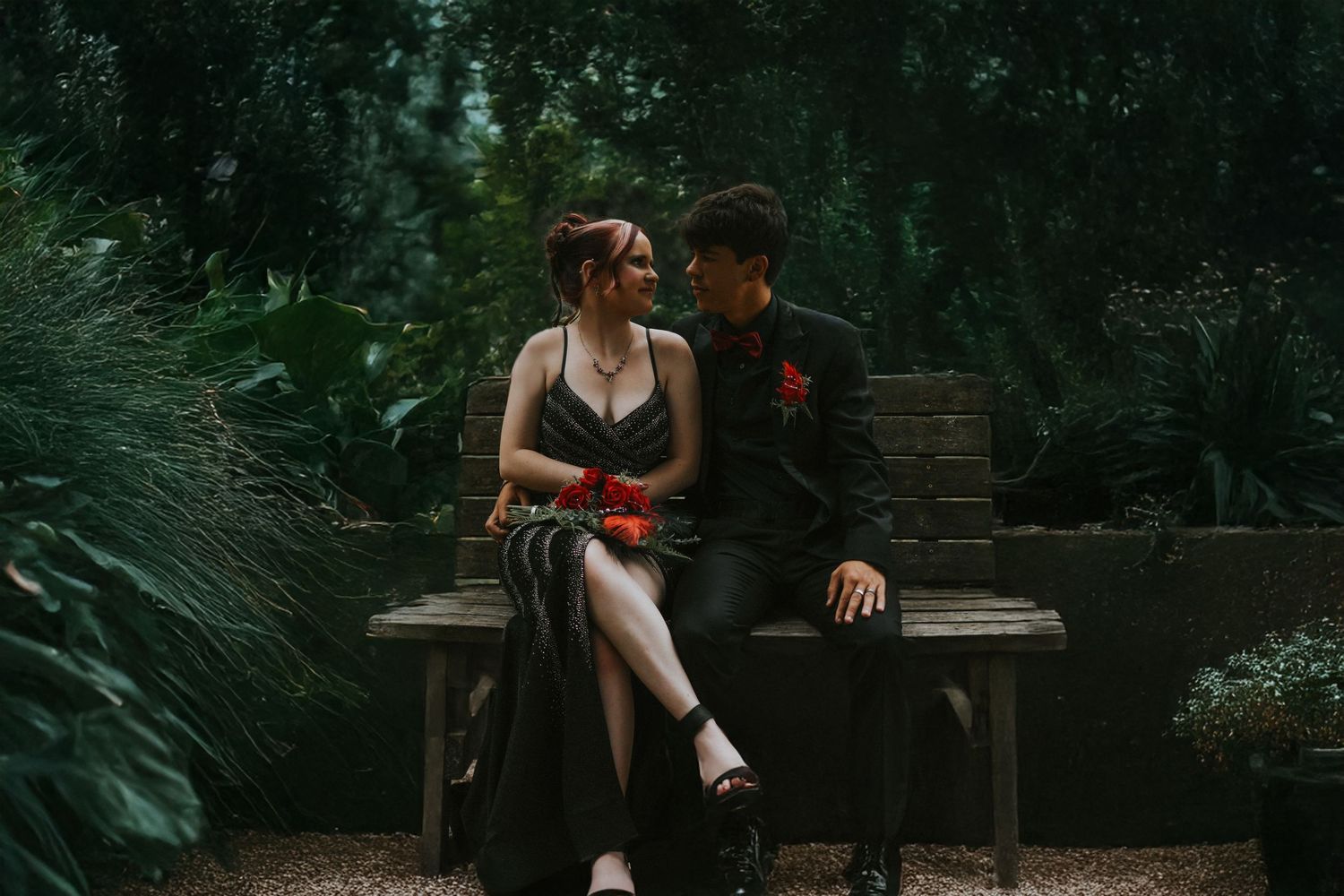 A couple in formal attire sits on a garden bench, gazing at each other romantically.