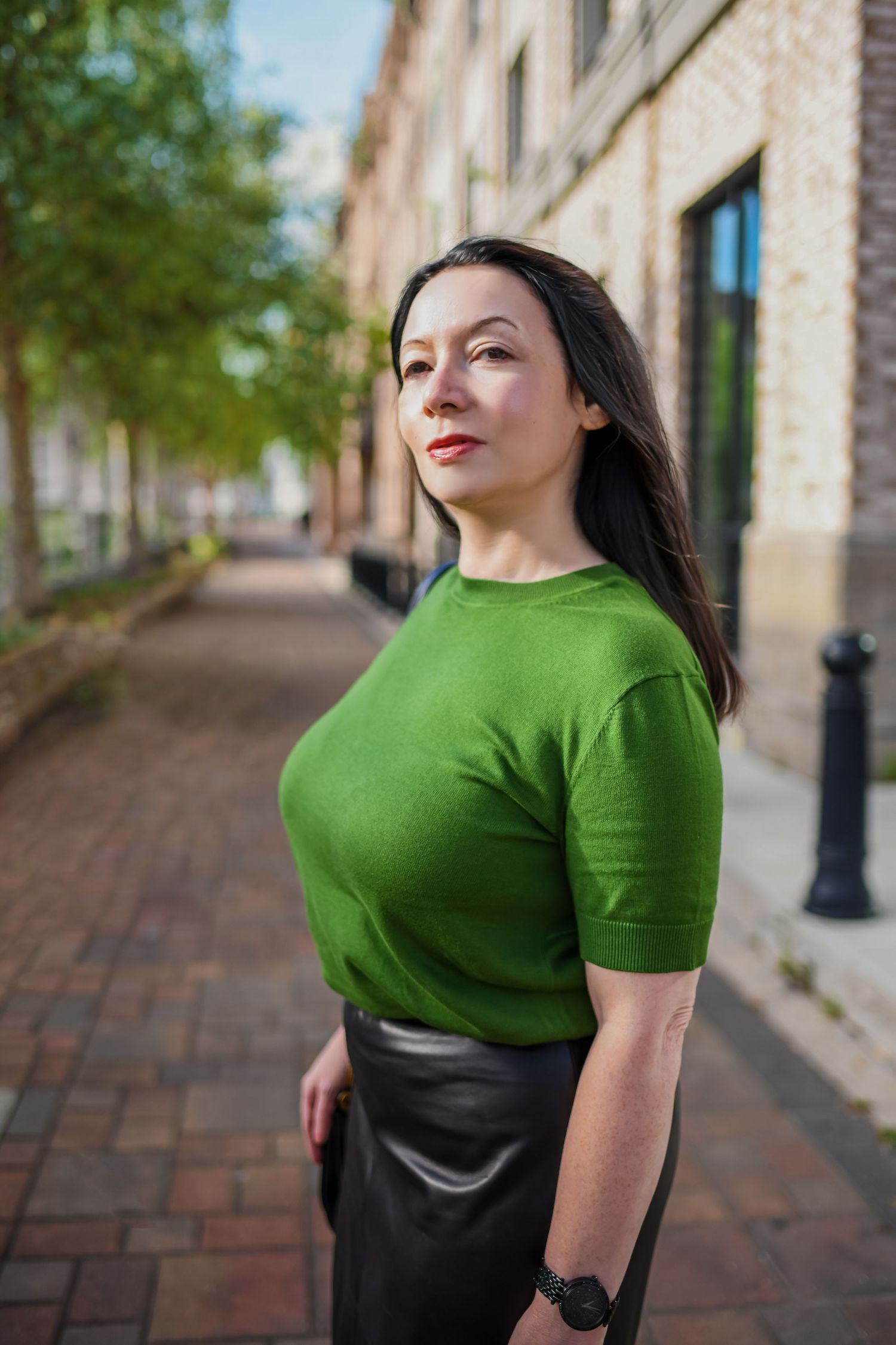 Woman in green top and black leather skirt poses on a brick sidewalk outside a historic building on a sunny day.