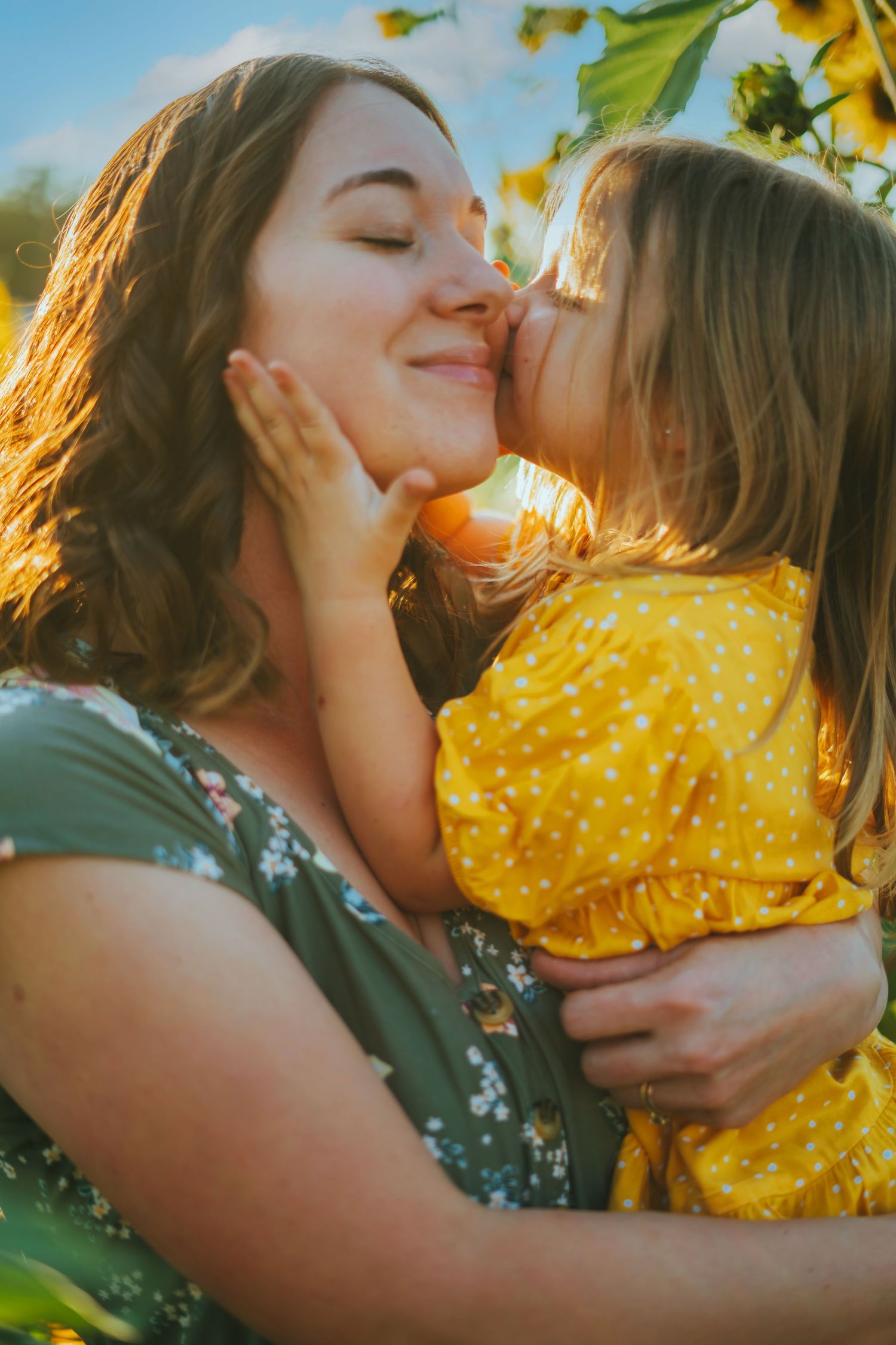 Joyful daughter kissing mothers cheek in sunflower field at golden hour sunset.