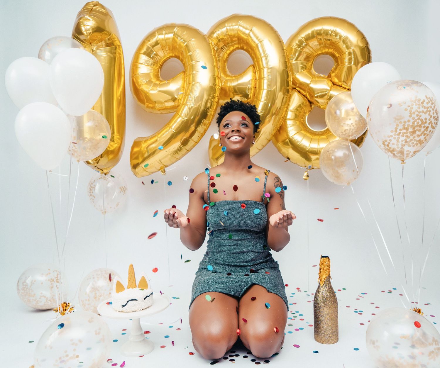 Woman celebrating 1998 birthday with gold balloons, confetti, cake, and champagne on white background.