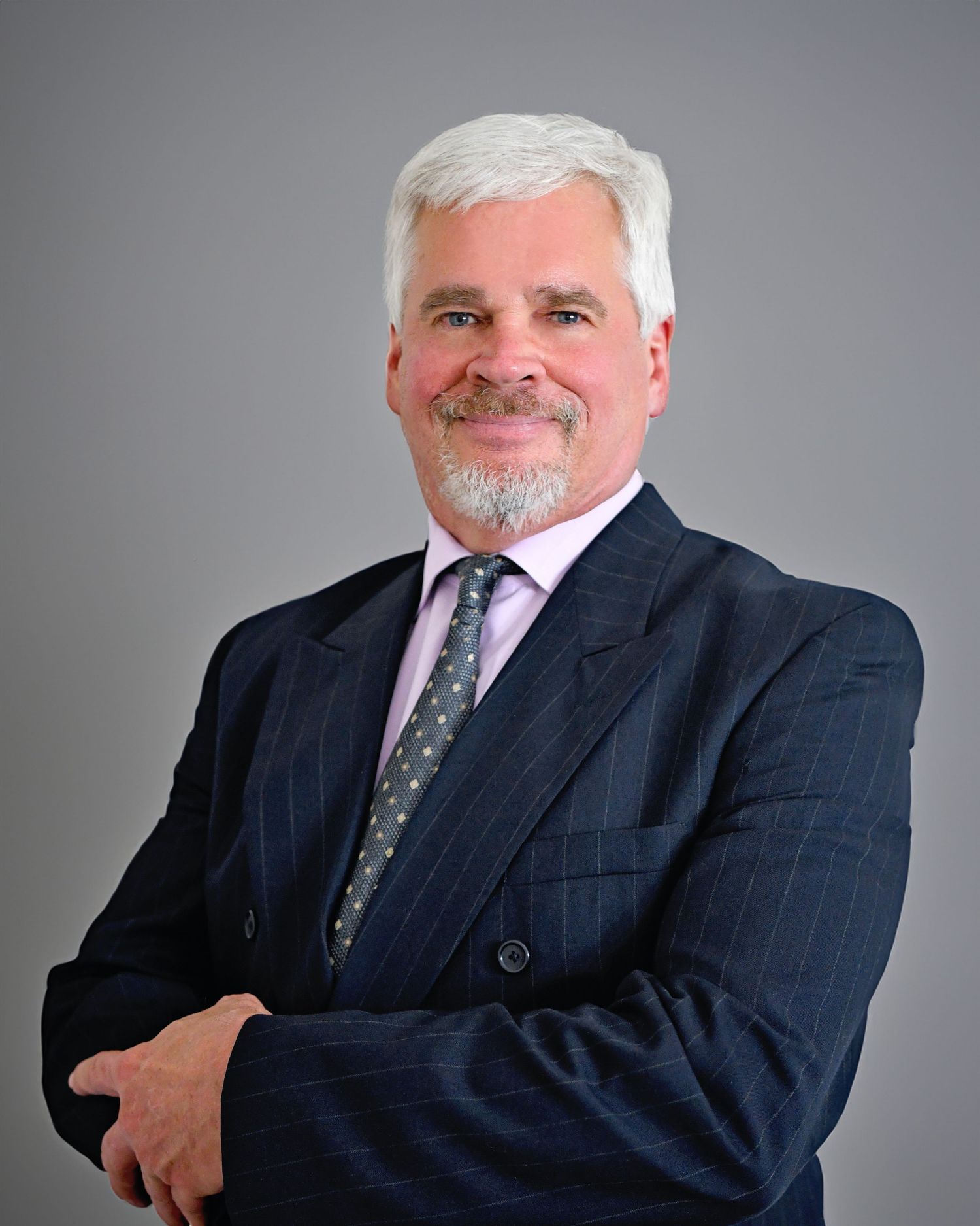 Smiling senior businessman with white hair and beard in navy pinstripe suit with arms crossed.