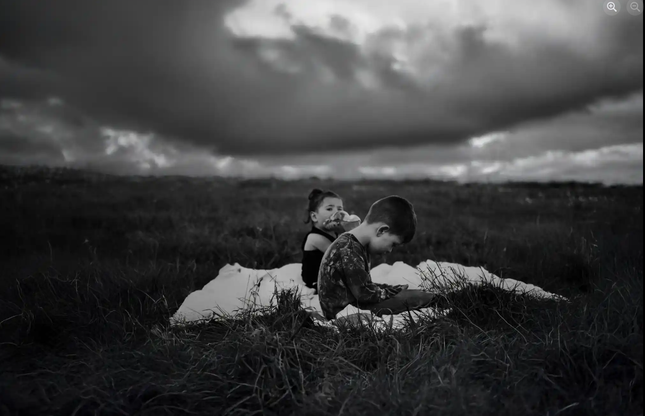 Black and white photo of two children lying in a grassy field under dramatic storm clouds.