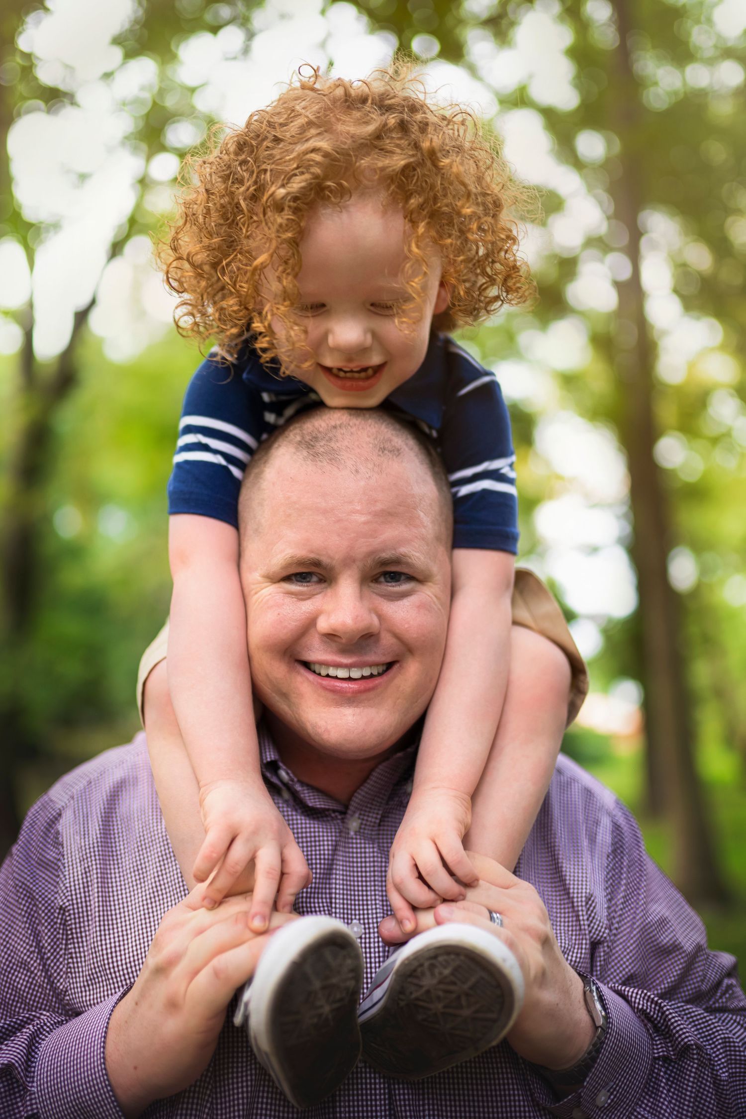 Smiling bald father with curly red-haired child sitting on his shoulders in a green park.