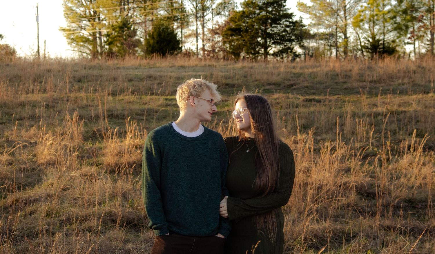 A young couple stands in a golden field at sunset, smiling at each other in a romantic outdoor portrait.