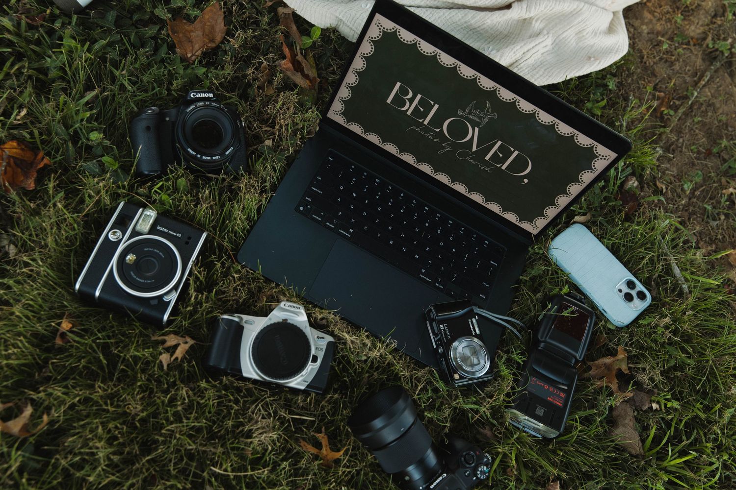Flat lay of Canon cameras, a smartphone, lens, and laptop displaying Beloved on green grass.