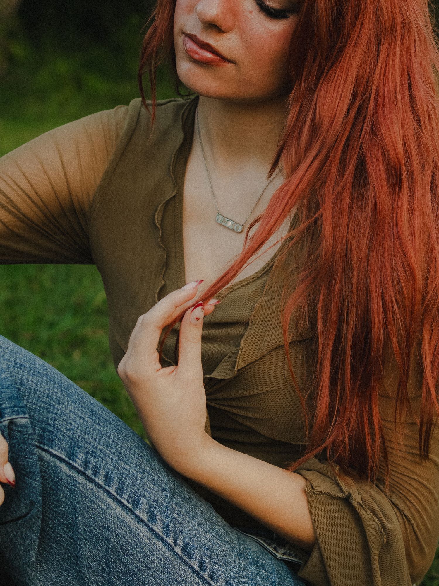 A young woman with long red hair wearing an olive top and jeans sits outdoors, gently touching her hair.