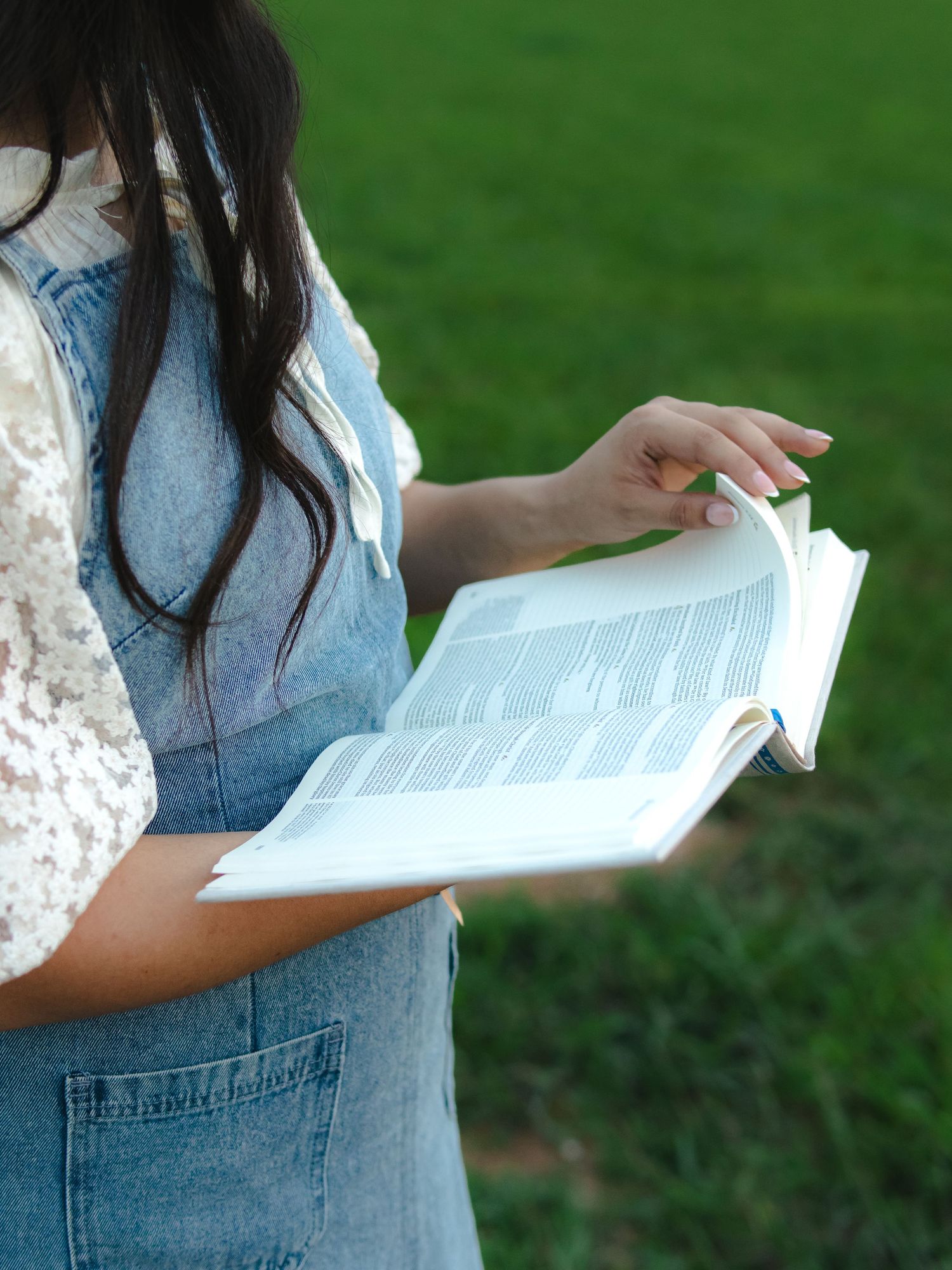 Young woman in denim overalls reading an open book outdoors on green grass background.